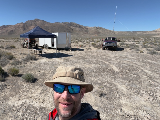 Selfie in the desert showing Jared, truck, trailer, side by side, antenna mast, radios, table, and chair